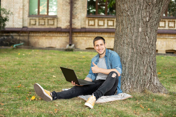 Portrait of positive man hold laptop show thumb up on grass at university campus. Business man sit on lawn work outdoors on computer in a park. Education and remote working concept.  - Photo, Image