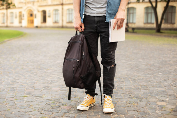 Man legs in yellow sneakers, wearing grey t-shirt, black skinny jeans at university campus in autumn. Front view of stylish man stand with book, waiting. Legs student in sneakers, standing on walkway. - Foto, afbeelding
