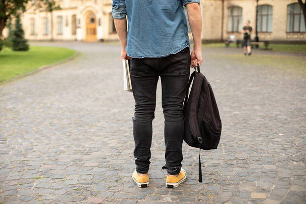 Man legs in yellow sneakers, denim shirt, black jeans at university campus. Back view of stylish man stand with book, waiting. Legs student in sneakers, standing on walkway. education concept - Фото, изображение