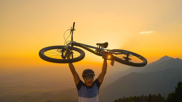 CLOSE UP, SUN FLARE: Happy Caucasian man shouts while holding his bike above his head at sunset. Cheerful male tourist celebrates winning a cross country bicycle race on a sunny summer evening. - Photo, Image