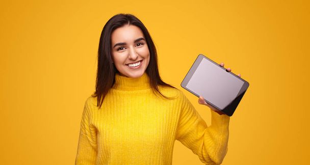 Positive young ethnic female model with long dark hair in yellow sweater smiling and looking at camera, while demonstrating tablet with black screen in bright studio - Photo, image