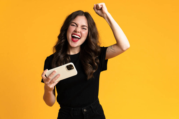 Weiße junge Frau macht Siegergeste, während sie ihr Handy isoliert auf gelbem Hintergrund benutzt - Foto, Bild