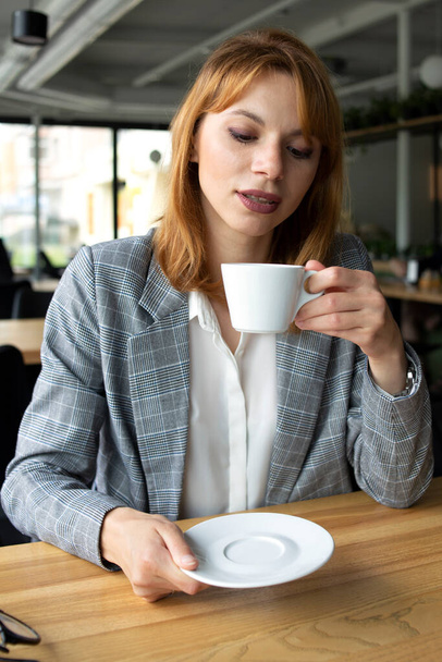Middle aged white woman with red hair in a grey jacket holding a white cup of coffee - Foto, afbeelding