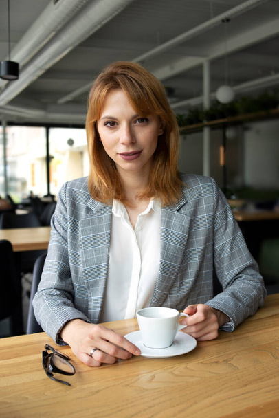 Seriöse Geschäftsfrau mit roten Haaren in grauer Jacke und weißem Hemd sitzt am Holztisch und hält eine Tasse Kaffee in der Hand, die gerade aussieht. - Foto, Bild