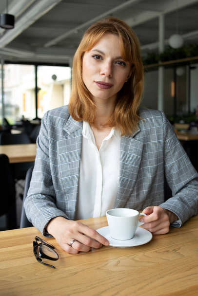 White middle aged white red hair woman at the wooden table with a cup of coffee and glasses wearing grey jacket and white shirt - Φωτογραφία, εικόνα