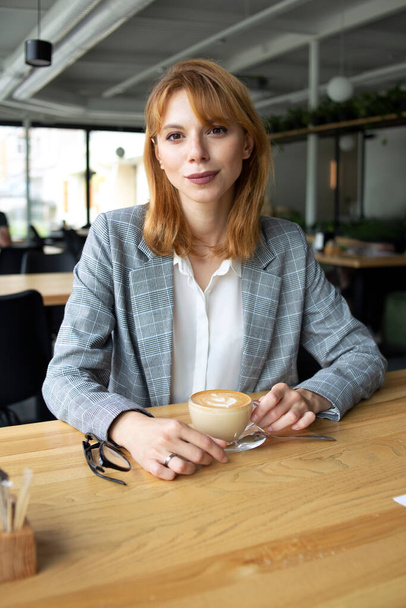 Slightly smiling young slim white woman with red hair wearing grey jacket and white shirt sitting at the wooden table and drinking coffee - 写真・画像
