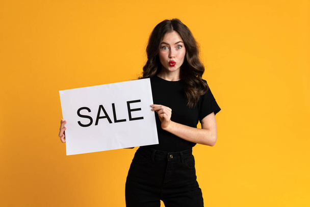 White woman expressing surprise while posing with sale placard isolated over yellow background - Foto, immagini