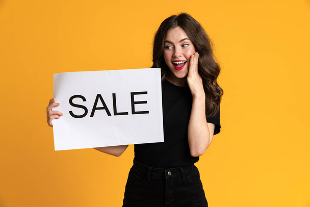 White young woman smiling while posing with sale placard isolated over yellow background - 写真・画像
