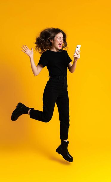 White young woman using mobile phone while gesturing and jumping isolated over yellow background - Zdjęcie, obraz