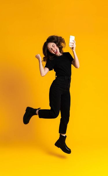 White young woman using mobile phone while gesturing and jumping isolated over yellow background - Foto, afbeelding