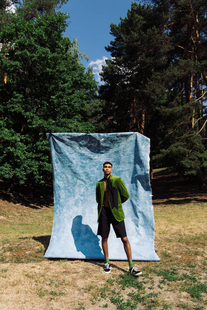 african american man in green blazer and black shorts standing with hands behind back near blue drape in park - Foto, afbeelding