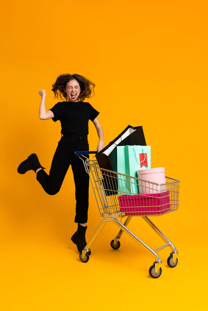 Excited woman making winner gesture while posing with shopping cart isolated over yellow background - Valokuva, kuva