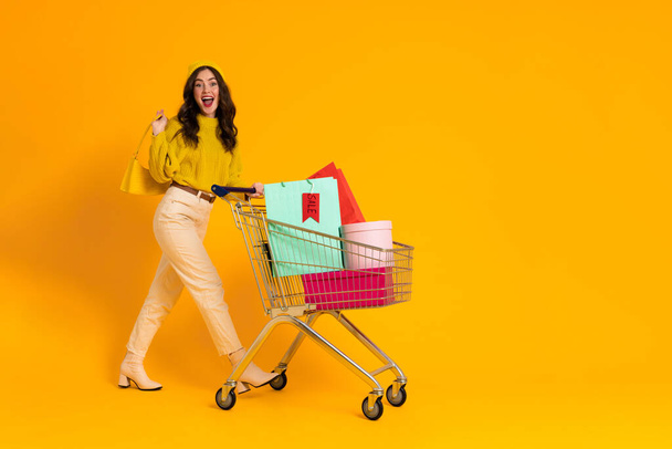 White excited woman laughing while posing with shopping cart isolated over yellow background - Photo, image