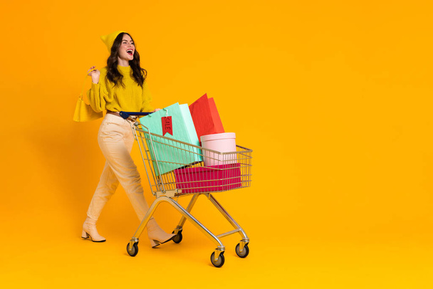 White excited woman laughing while posing with shopping cart isolated over yellow background - Foto, immagini