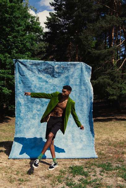 trendy african american man in velvet blazer and shorts pointing with finger near blue drape in forest - Foto, imagen