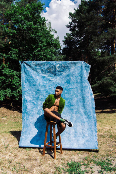 full length of african american man in black shorts and green blazer posing on stool in forest near blue cloth - Foto, afbeelding