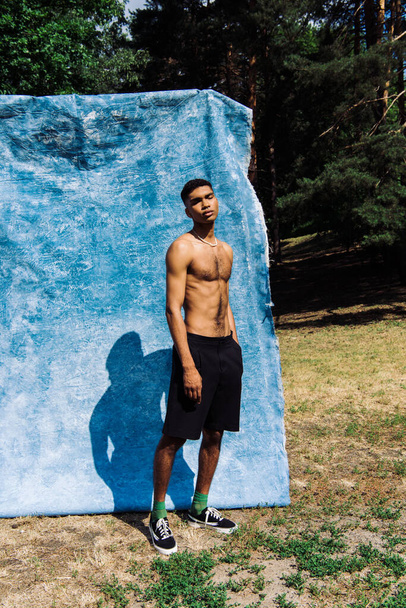 full length of shirtless african american man posing with hand in pocket of black shorts near blue drapery in park - Φωτογραφία, εικόνα