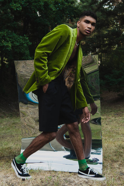 full length of african american man in green blazer and hand in pocket of black short walking near mirror in park - Foto, Imagen
