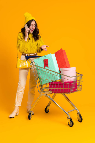 Excited woman talking on cellphone while posing with shopping cart isolated over yellow background - Foto, Imagen