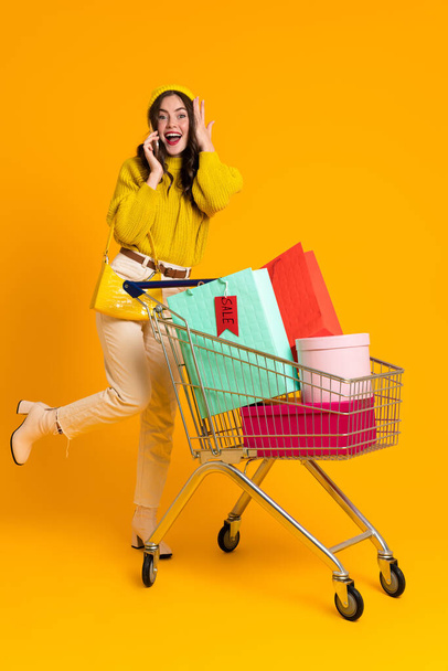 Excited woman talking on cellphone while posing with shopping cart isolated over yellow background - Foto, Imagem