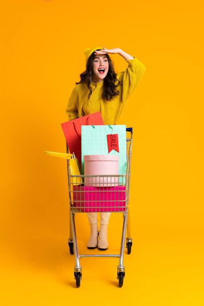 White excited woman gesturing while posing with shopping cart isolated over yellow background - Фото, зображення