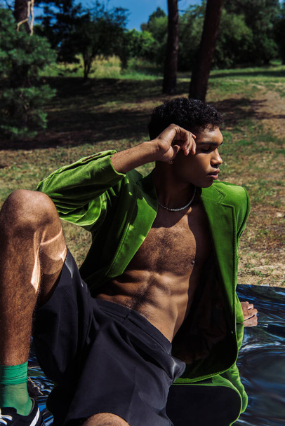 african american man with closed eyes sitting in green blazer on mirror surface in park - Φωτογραφία, εικόνα