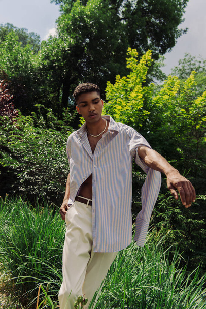 trendy african american man in blue shirt posing with hand in pocket in park - Fotó, kép