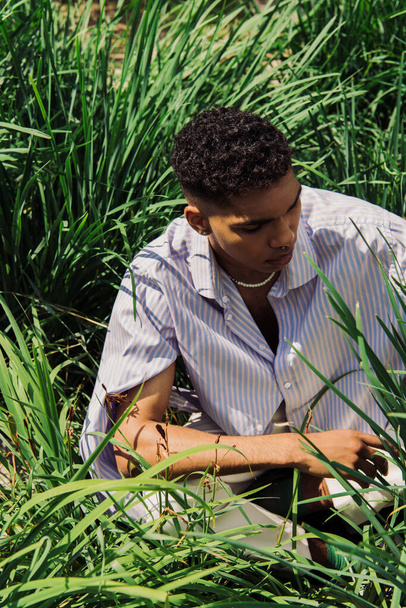 high angle view of african american man in fashionable shirt sitting on green grass in park - Foto, immagini