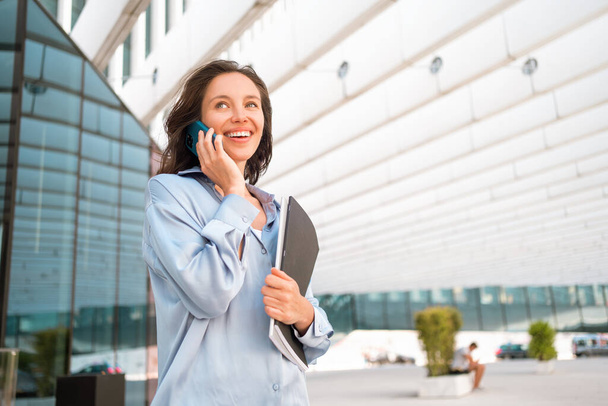 Attractive brunette businesswoman talking mobile phone with client. Pretty caucasian female business person smile when phone conversation. Woman managing business talking via mobile phone standing - Foto, afbeelding