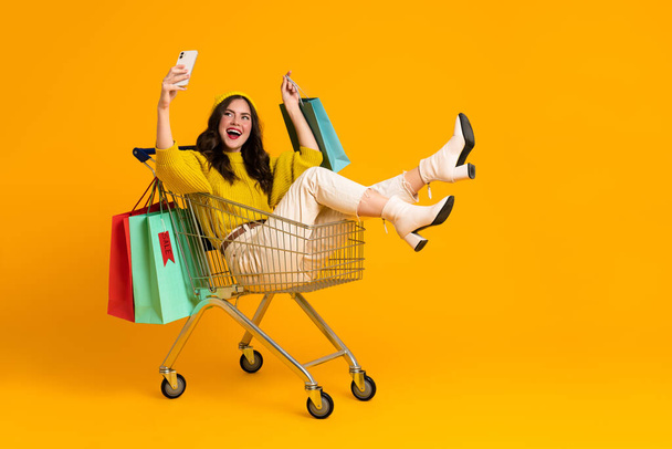 White excited woman taking selfie on cellphone in shopping cart isolated over yellow background - Foto, immagini