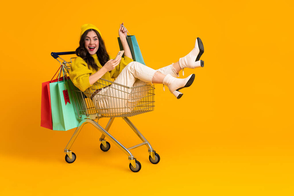 White excited woman laughing and using cellphone in shopping cart isolated over yellow background - Φωτογραφία, εικόνα