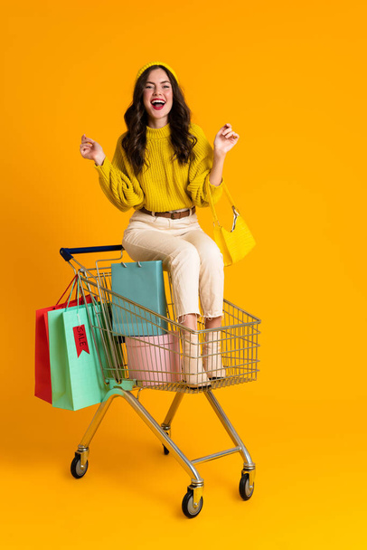 White woman laughing while sitting in shopping cart isolated over yellow background - Foto, Imagen