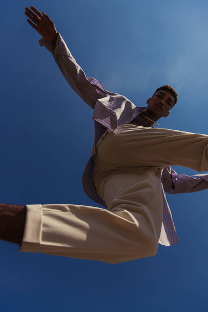 bottom view of african american man in white pants jumping under blue sky - Foto, Imagen