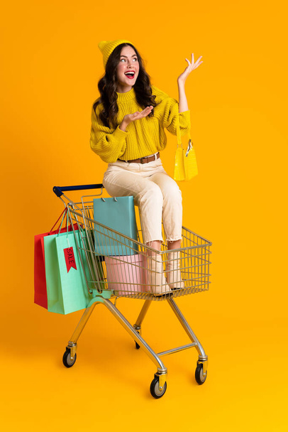 White woman holding copyspace while sitting in shopping cart isolated over yellow background - Zdjęcie, obraz