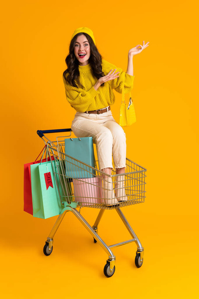 White woman holding copyspace while sitting in shopping cart isolated over yellow background - Fotografie, Obrázek