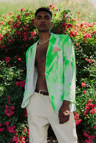 young african american man in stylish blazer and beads standing near blossoming bushes - 写真・画像