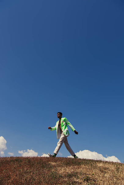side view of african american man in blazer and pants walking in field under clear sky - Φωτογραφία, εικόνα