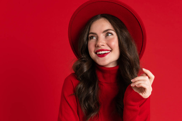 witte jonge vrouw dragen hoed kijken en glimlachen opzij geïsoleerd over rode achtergrond - Foto, afbeelding