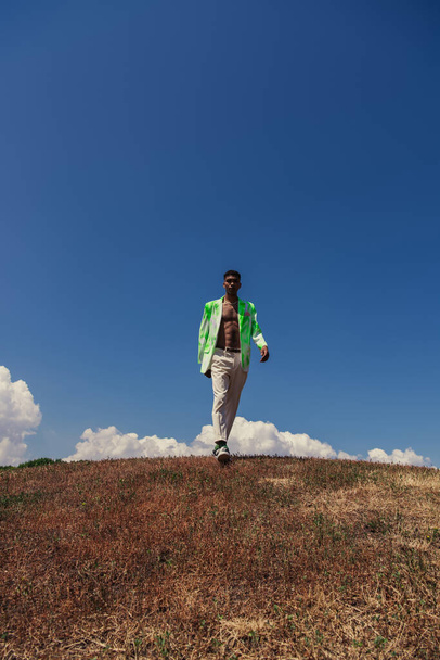 full length of african american man in trousers and blazer walking in field under blue sky with white clouds - Φωτογραφία, εικόνα