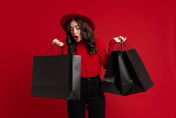 White woman expressing surprise while posing with shopping bags isolated over red background - Fotografie, Obrázek