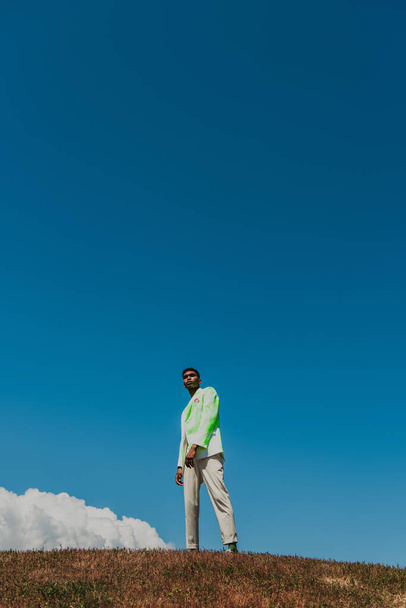 african american man in stylish blazer and trousers standing in meadow under blue sky - Foto, Imagen