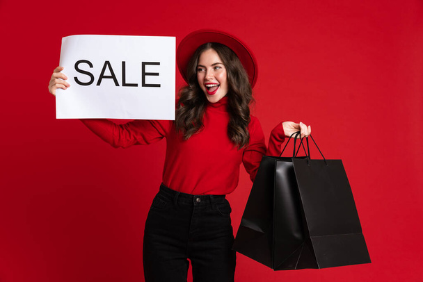 White woman smiling while posing with shopping bags and sale placard isolated over red background - Photo, Image