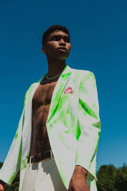 low angle view of african american man in white and green blazer standing under blue sky and looking away - Foto, Imagen