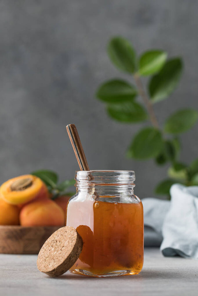 A jar with apricot jam and a spoon close-up on a gray background. In the background is a plate with fresh apricots and a branch with green leaves. Fruit preservation. - Foto, afbeelding