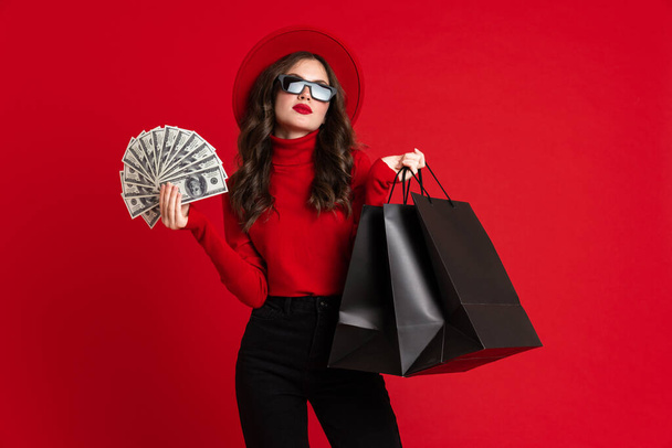 White woman in sunglasses posing with shopping bags and dollars isolated over red background - Foto, afbeelding