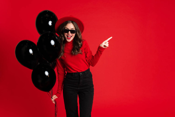 White woman pointing finger aside while posing with black balloons isolated over red background - Fotoğraf, Görsel