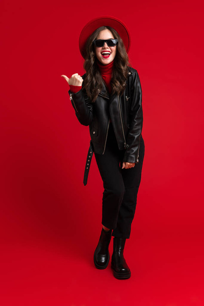 White woman in leather jacket smiling and pointing finger aside isolated over red background - Foto, Bild