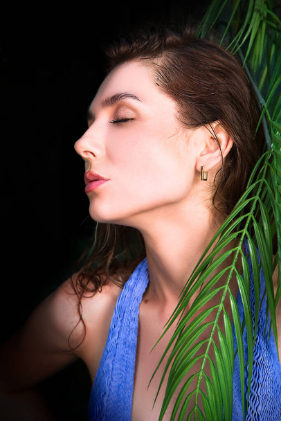 Portrait of a young woman in water drops near a palm tree on a black background. - Foto, imagen