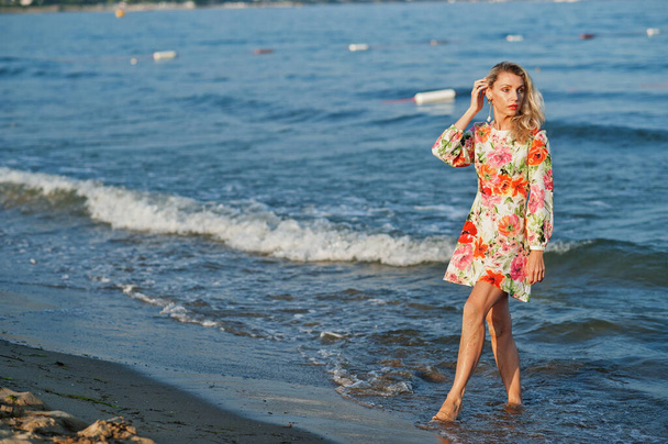 Blonde woman wearing dress standing on sand beach at sea shore enjoying view of sunset. - Foto, imagen