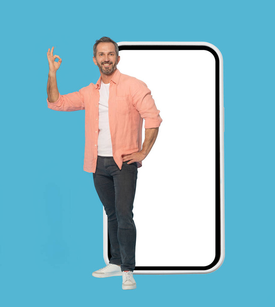 Gesturing OK standing in front big smartphone with white screen handsome middle aged grey haired man smiling wearing peach shirt isolated on blue background. Mobile app advertising mock up.  - Valokuva, kuva